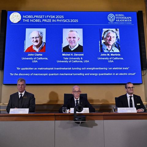 Drei weiße, ältere Männer in Anzügen verkünden die Empfänger des Nobelpreises für Physik 2025
