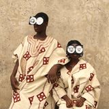 Zwei junge Menschen posieren vor einer rauen Wand, in reich bestickten Gewändern aus Aso Oke – jenem handgewebten Stoff aus Nigeria, der traditionell bei Zeremonien getragen wird. Ihre Gesichter sind größtenteils von futuristischen Brillen aus recycelten VHS-Kassetten verdeckt – eine witzige und prägnante Metapher für die Verwandlung veralteter Technik in ein neues Ritualobjekt und Werkzeug des kulturellen Ausdrucks