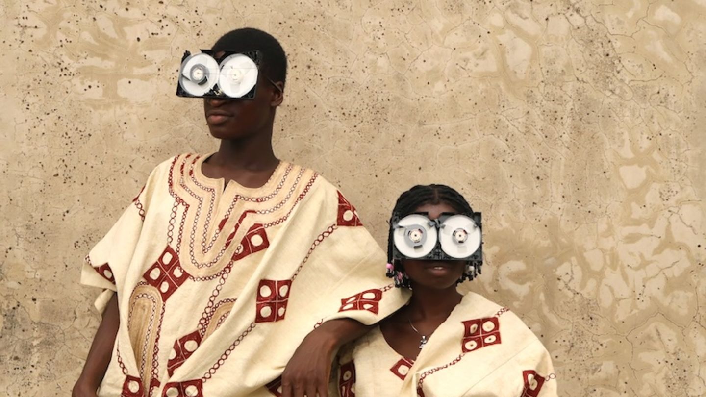 Zwei junge Menschen posieren vor einer rauen Wand, in reich bestickten Gewändern aus Aso Oke – jenem handgewebten Stoff aus Nigeria, der traditionell bei Zeremonien getragen wird. Ihre Gesichter sind größtenteils von futuristischen Brillen aus recycelten VHS-Kassetten verdeckt – eine witzige und prägnante Metapher für die Verwandlung veralteter Technik in ein neues Ritualobjekt und Werkzeug des kulturellen Ausdrucks