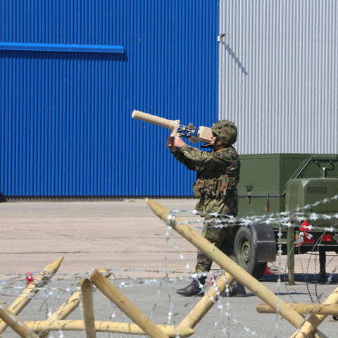 Ein maskierter Bundeswehrsoldat zielt mit eine Art Strahlenkanone auf eine rote Drohne, die vor ihm fliegt
