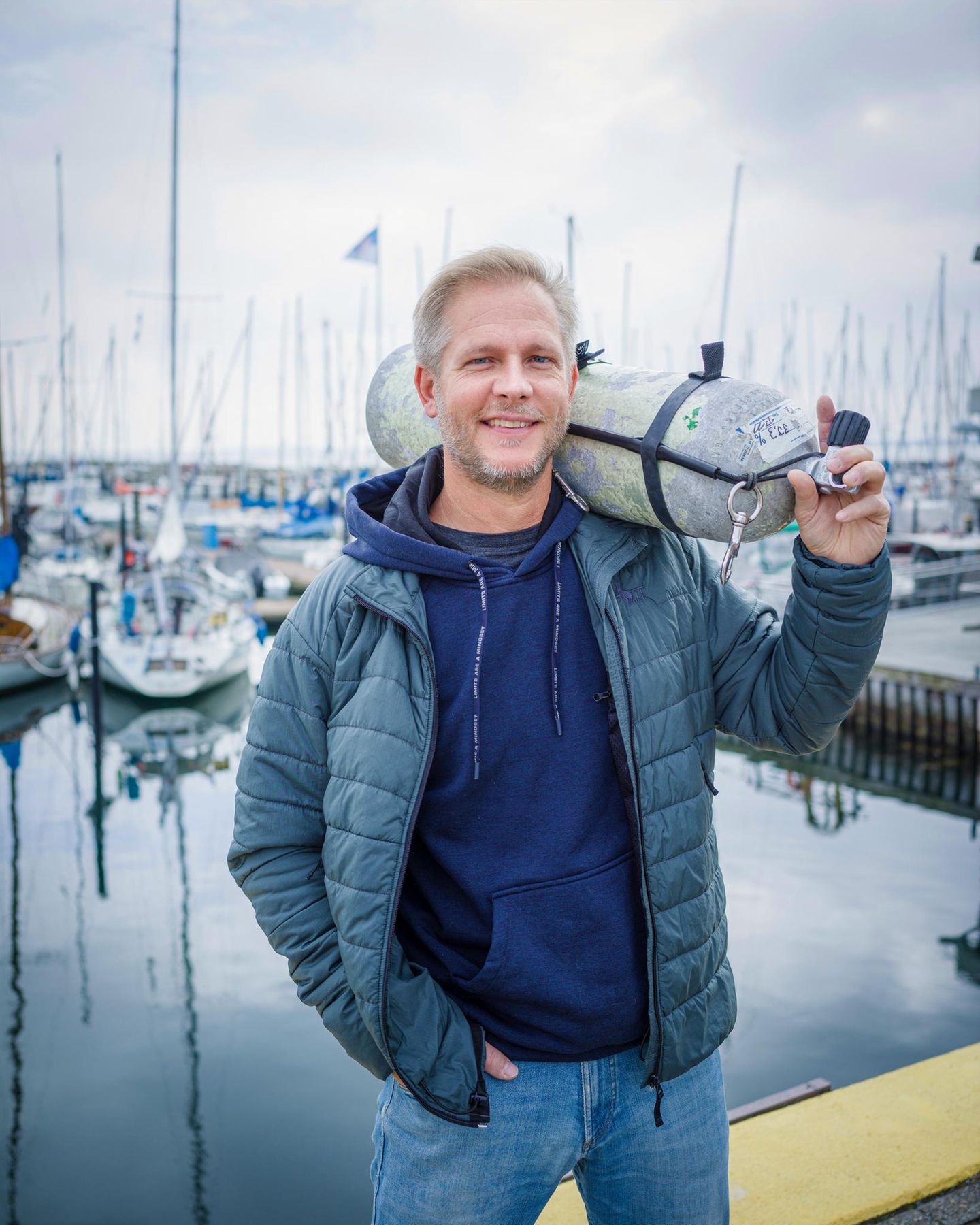 Der Forschungstaucher Florian Huber steht mit einer Druckluftflasche auf der Schulter in einen Yachthafen
