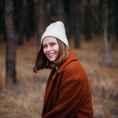 Eine Frau steht im Wald. Sie lächelt in die Kamera.