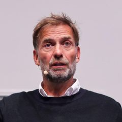 Jürgen Klopp sitzt auf einer Bühne und gestikuliert beim Sprechen mit erhobener rechter Hand