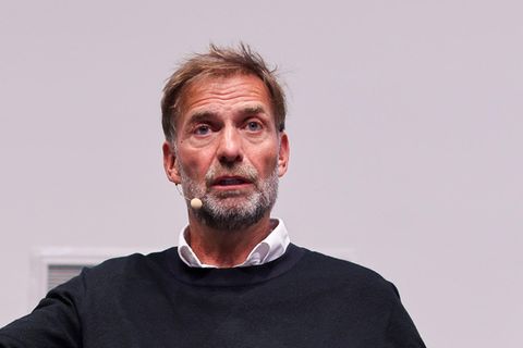 Jürgen Klopp sitzt auf einer Bühne und gestikuliert beim Sprechen mit erhobener rechter Hand