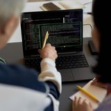 Ein älterer Herr zeigt auf einen Laptop, auf dem Codereihen laufen