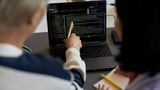 Ein älterer Herr zeigt auf einen Laptop, auf dem Codereihen laufen