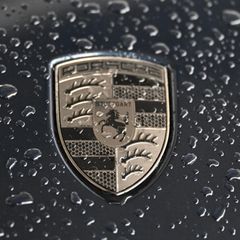 Das Logo von Porsche glänzt golden auf einer schwarzen Motorhaube voller Regentropfen