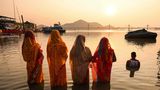 Vier in traditioneller Kleidung bekleidete Inderinnen stehen bis über ihre Knie im Wasser des Brahmaputra und beten die Sonne an