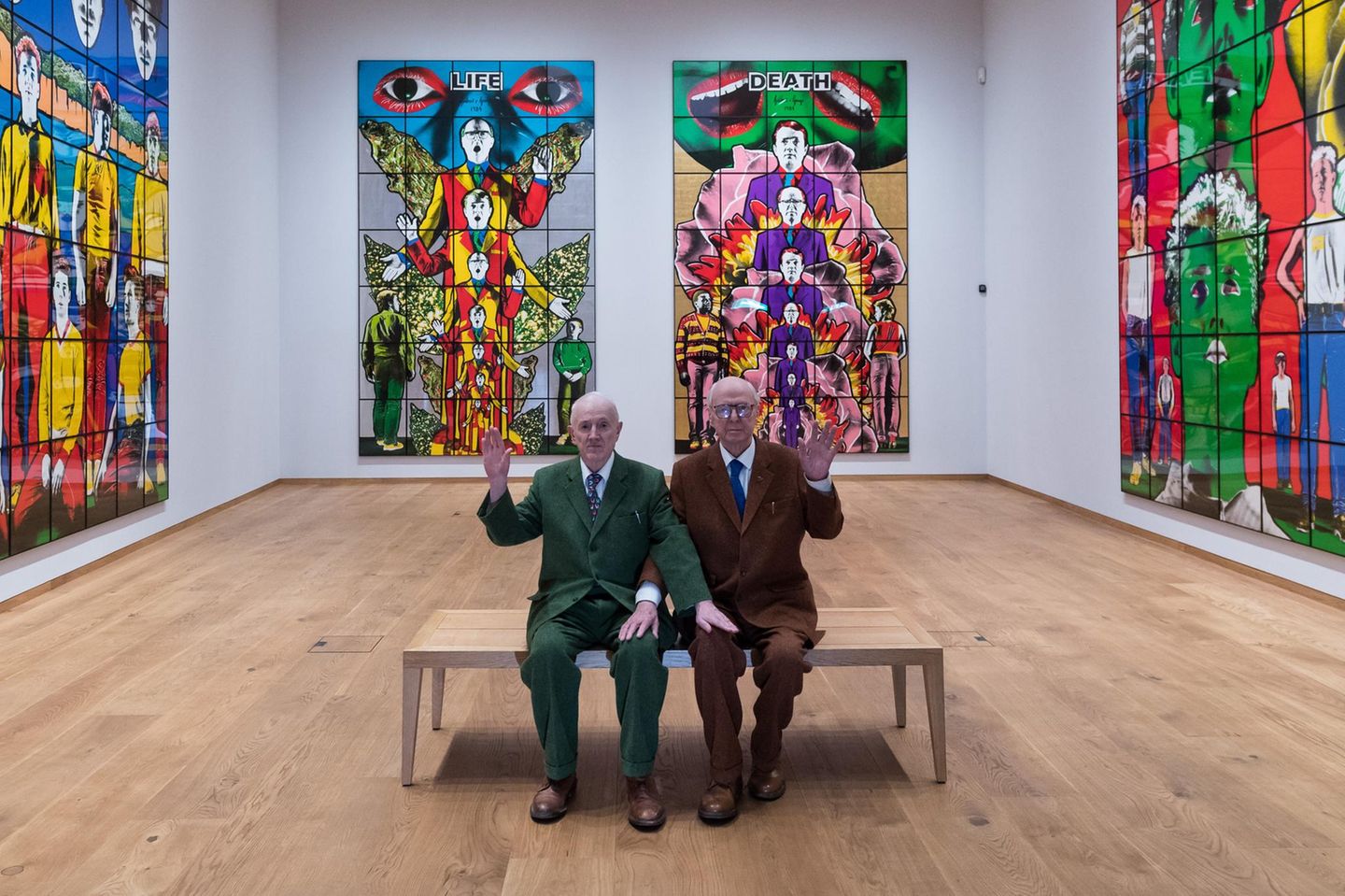 Gilbert und George sitzen auf einer Bank in ihrer Ausstellung und winken, 2025