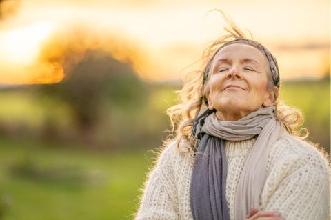 Eine Frau genießt beim Spazieren das Wetter, freut sich über die Rentenerhöhung, im Hintergrund unscharf die Sonne hinter Bäumen