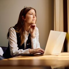 Symbolbild Unternehmensberaterin: Eine junge Frau sitzt am Laptop und schaut aus dem Fenster