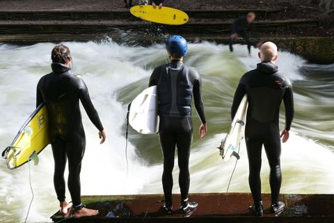 Vier Surfer stehen in Neoprenanzügen und mit ihren Boards unterm Arm an der Eisbachwelle in München
