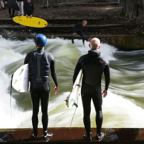 Vier Surfer stehen in Neoprenanzügen und mit ihren Boards unterm Arm an der Eisbachwelle in München