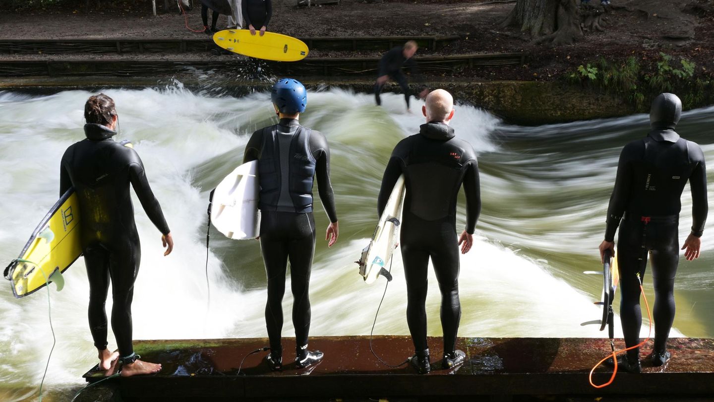 Vier Surfer stehen in Neoprenanzügen und mit ihren Boards unterm Arm an der Eisbachwelle in München