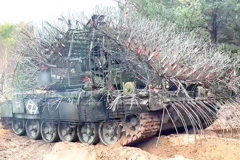 Ein russischer, mit abstehenden Drahtseilen verkleideter Panzer rollt über eine ländliche Straße.