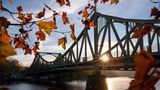 Potsdam, Deutschland. Ein Novembermorgen taucht die Glienicker Brücke in warmen Sonnenstrahlen. Orangefarbene und gelbe Blätter umrahmen dieses historische Symbol für deutsche Teilung und Wiedervereinigung