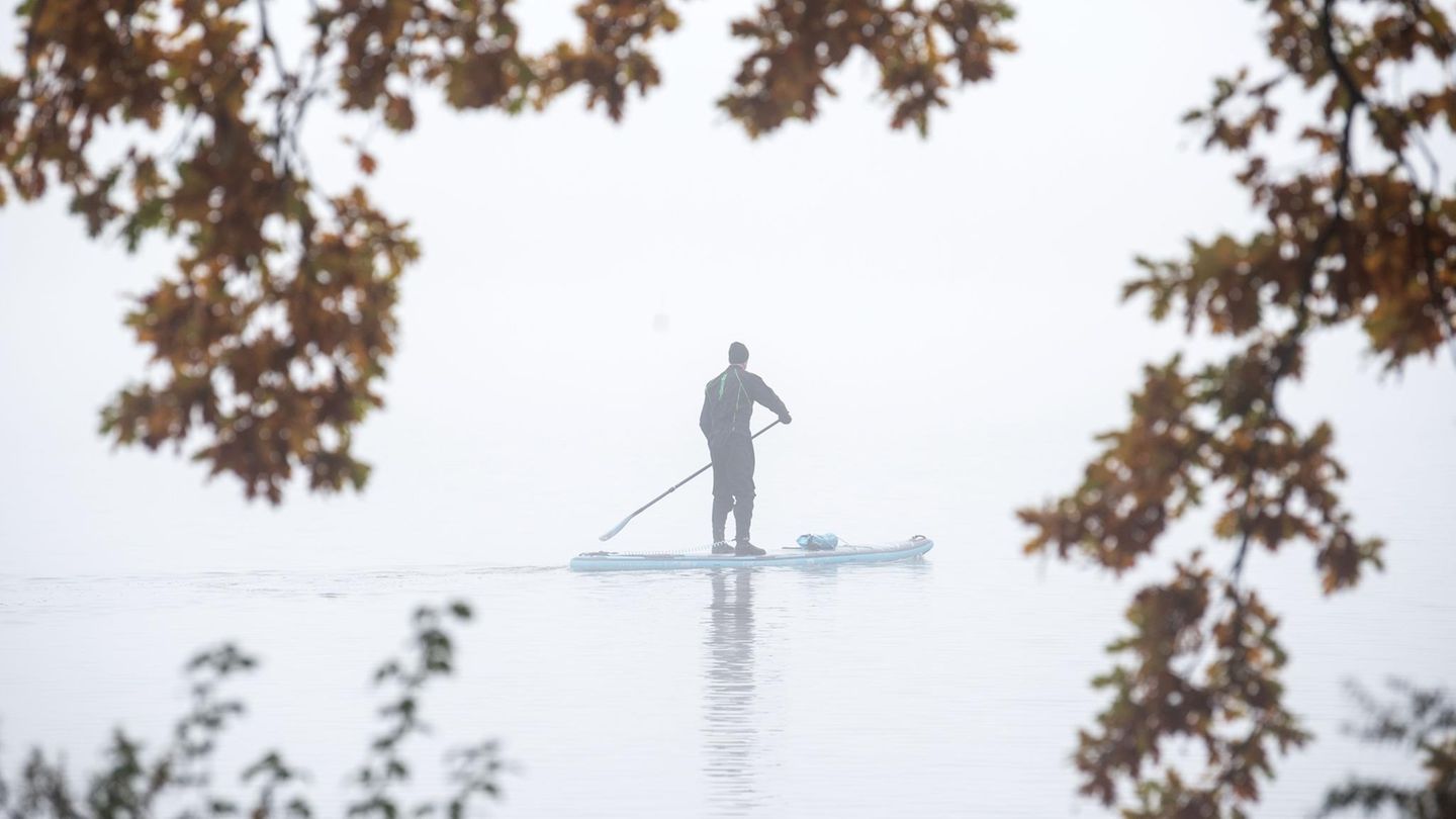 Hamburg, Deutschland. Inmitten nebelverhangener Ruhe paddelt ein Mann mit einem Stand-up-Paddleboard konzentriert auf der Außenalster an herbstlich verfärbten Bäumen entlang