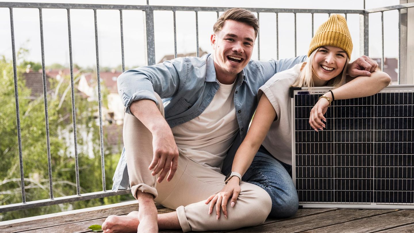 Zwei junge Personen sitzen auf dem Boden eines Balkons und lachen in die Kamera, die Rechte lehnt sich auf ein Solarpanel
