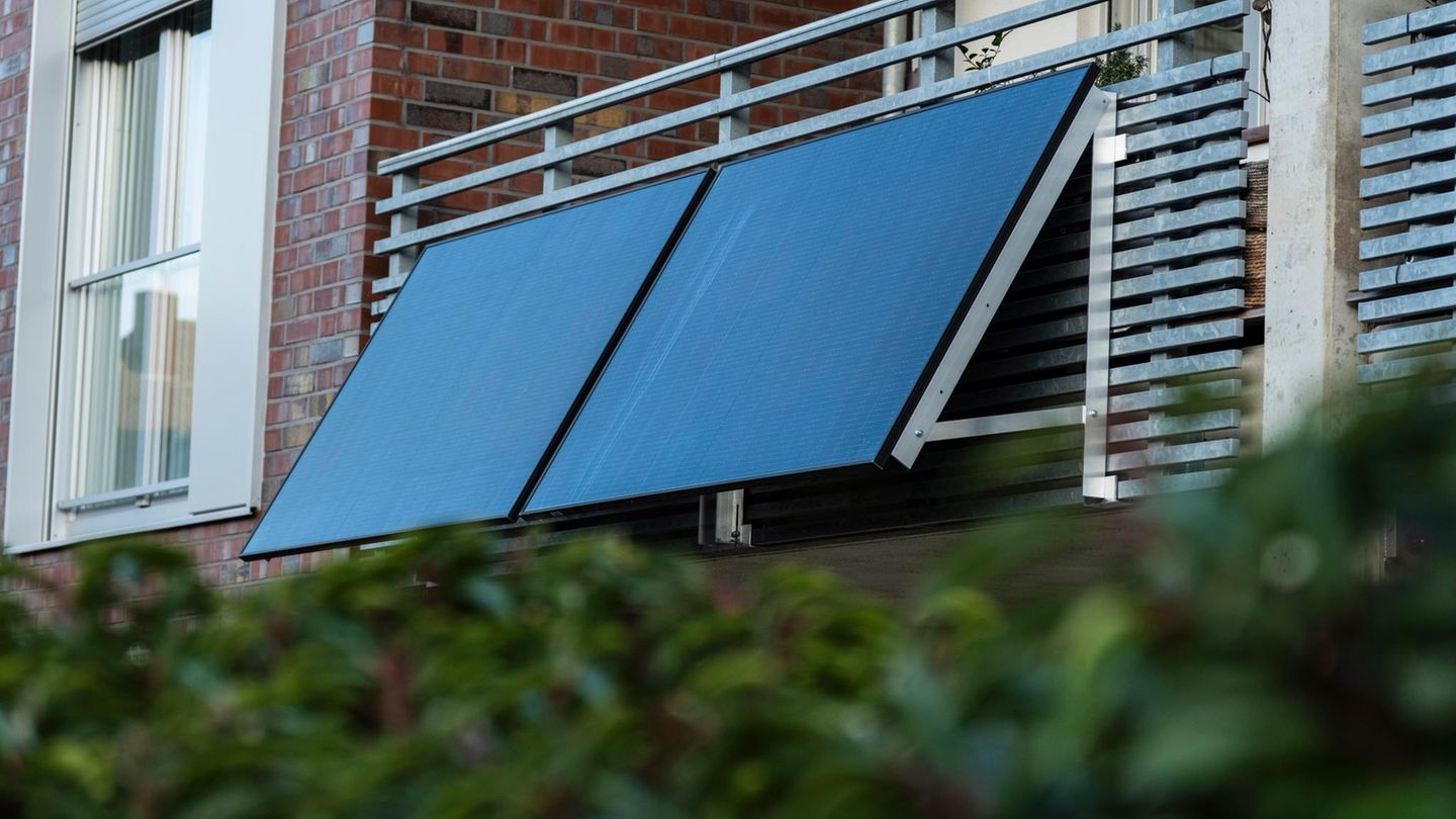 Zwei Solarmodule hängen an einem Gitterbalkon, davor ist eine grüne Hecke zu sehen.