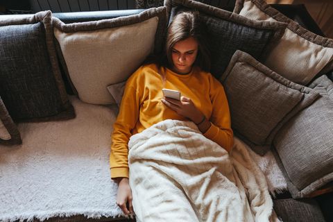 Eine Frau schaut auf ihr Handy und sitzt auf dem Sofa (Symbolbild Prokrastination)