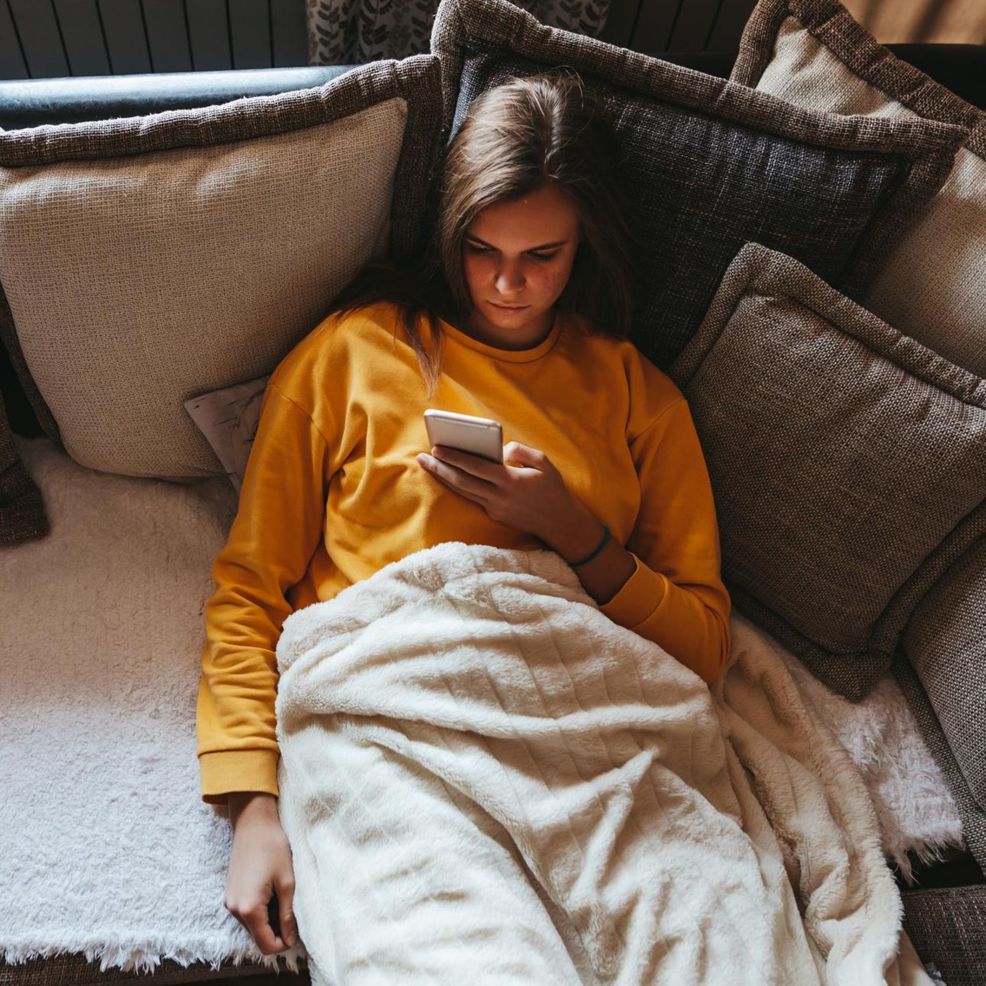 Eine Frau schaut auf ihr Handy und sitzt auf dem Sofa (Symbolbild Prokrastination)