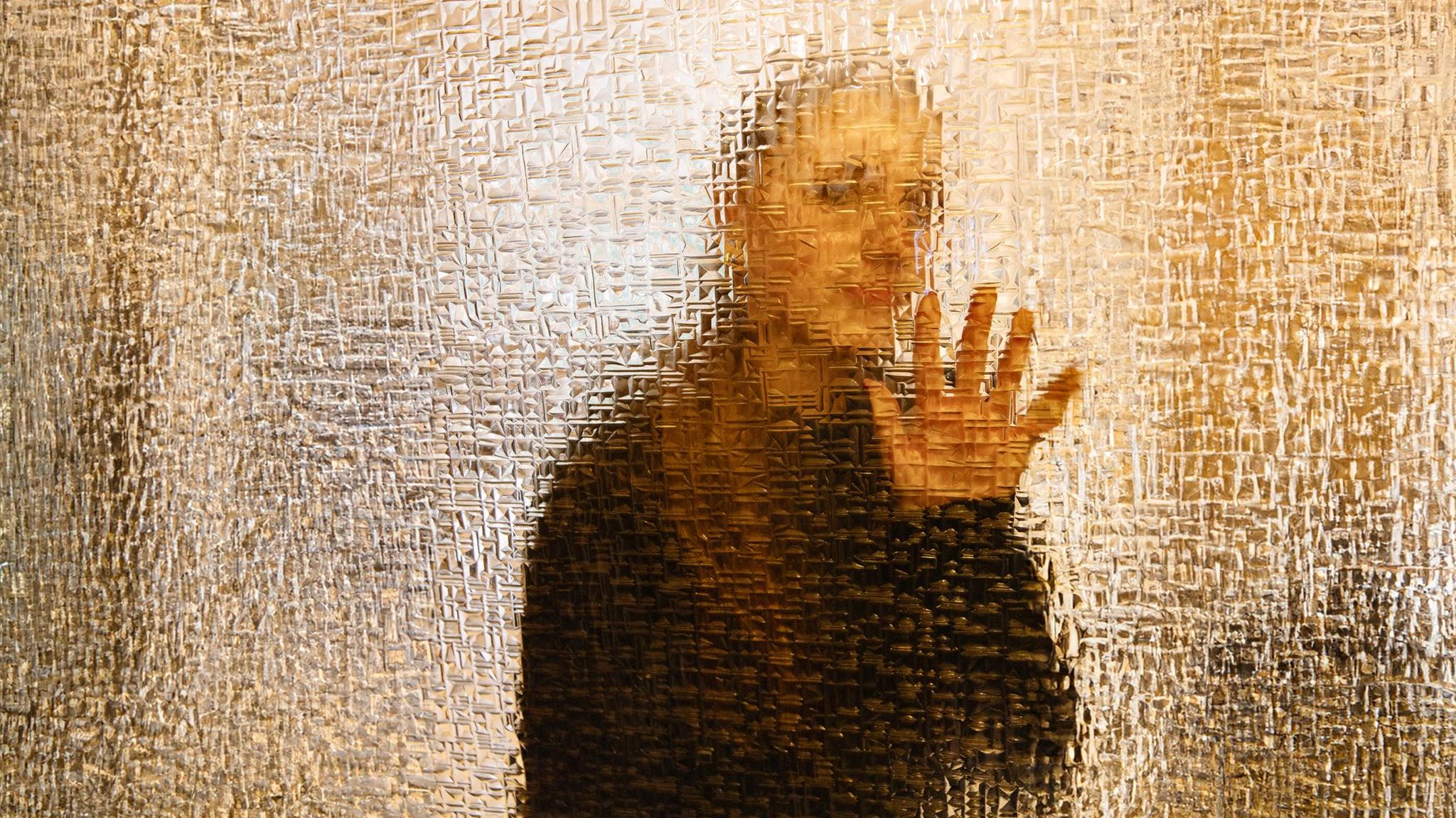 Hinter einer Glaswand erkennt man unscharf eine weibliche Gestalt. Hadert sie mit ihrewr identität?