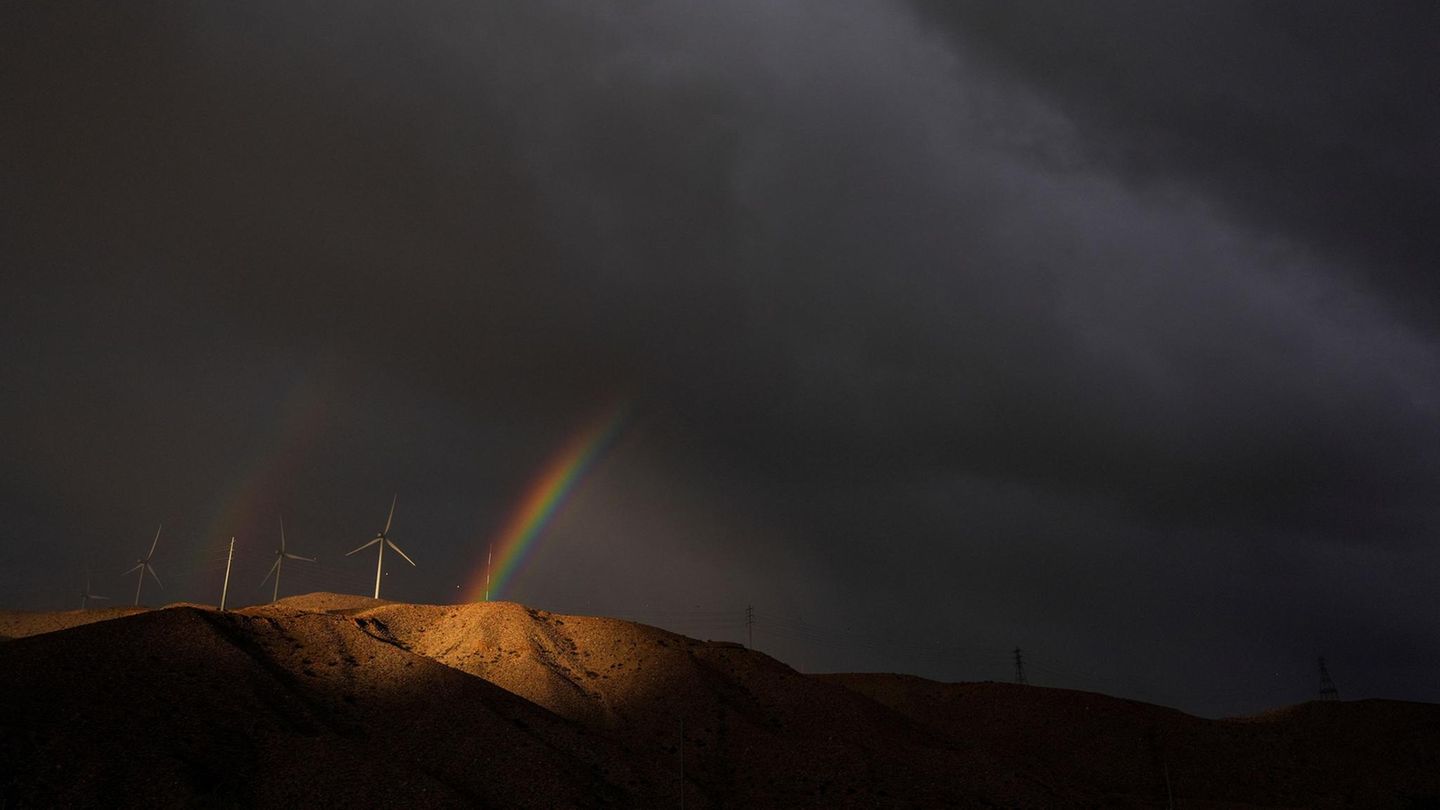 Cathedral City, USA. Mystischer Anblick: Mitten in dunklen Gewitterwolken erscheint ein doppelter Regenbogen hinter Windturbinen auf einem Hügel