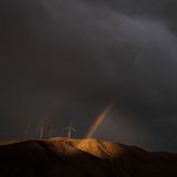 Cathedral City, USA. Mystischer Anblick: Mitten in dunklen Gewitterwolken erscheint ein doppelter Regenbogen hinter Windturbinen auf einem Hügel
