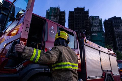 In Hongkong steigt ein Feuerwehrmann aus einem Fahrzeug, während im Hintergrund vier Wohn-Hochhäuser rauchen