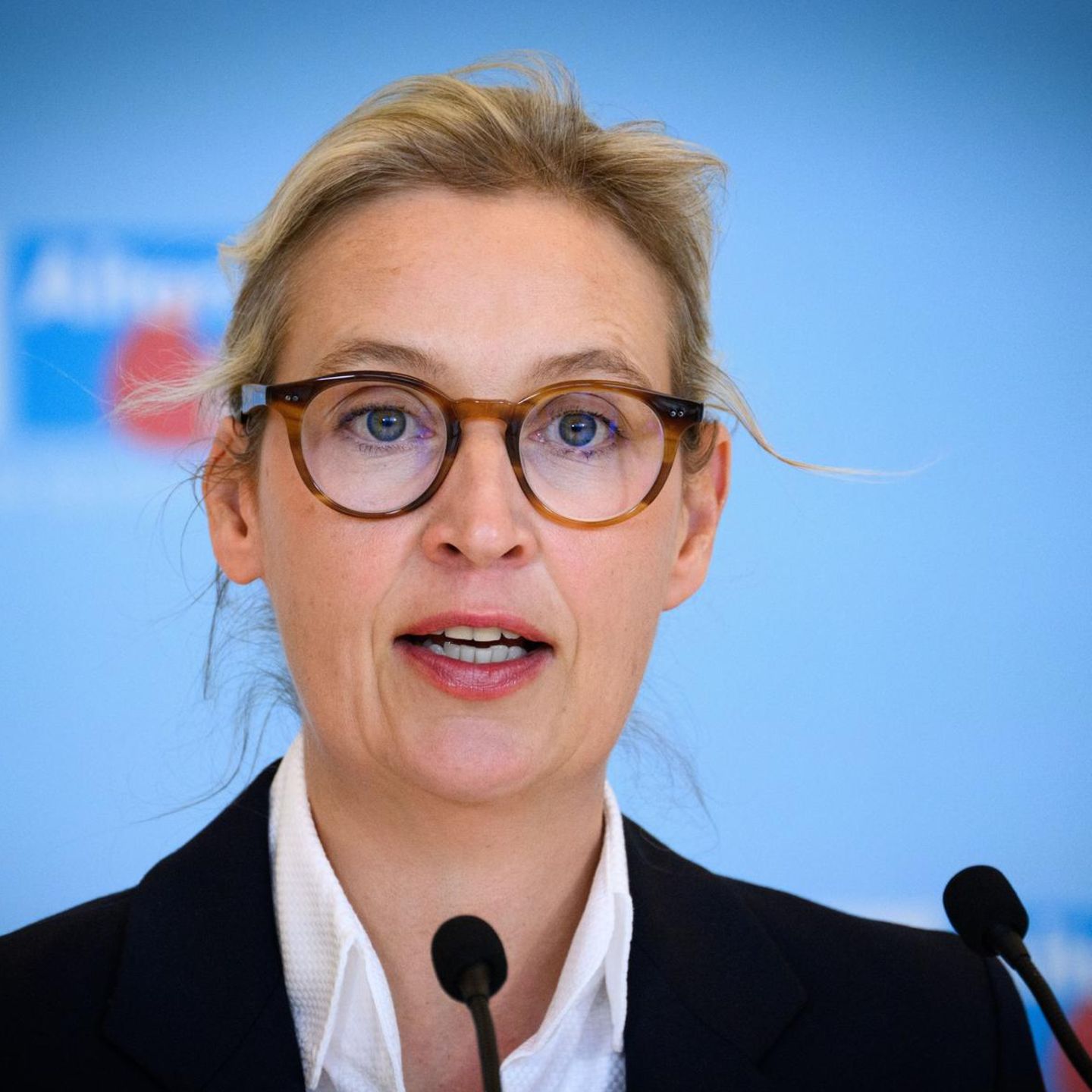 Alice Weidel spricht vor einer blauen Wand mit AfD-Logo in zwei Mikrofone