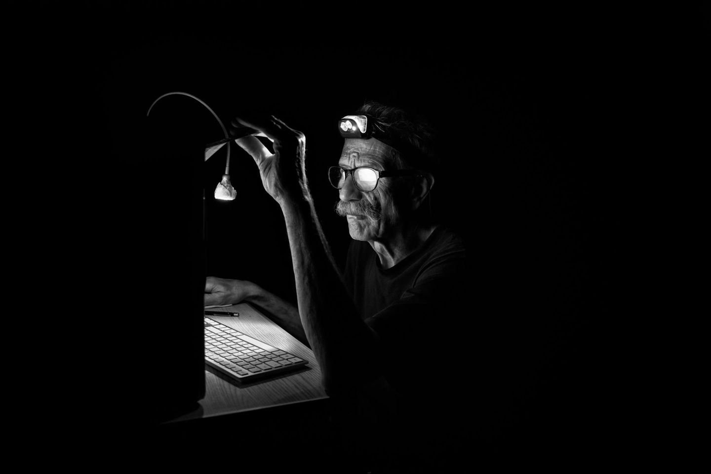 Eine Person sitzt im Dunklen vor einem Computer und richtet die Schreibtischlampe aus, um besser sehen zu können