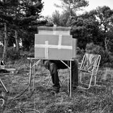 Eine Person sitzt im Garten hinter einem Karton, welcher auf einem Tisch steht, sodass nur seine Beine rausgucken