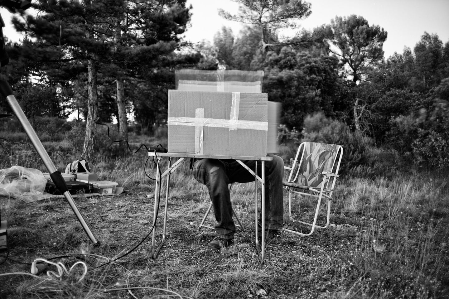 Eine Person sitzt im Garten hinter einem Karton, welcher auf einem Tisch steht, sodass nur seine Beine rausgucken