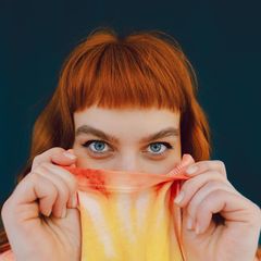 Eine junge Frau versteckt ihre untere Gesichtshälfte in ihrem T-shirt und schaut in die Kamera