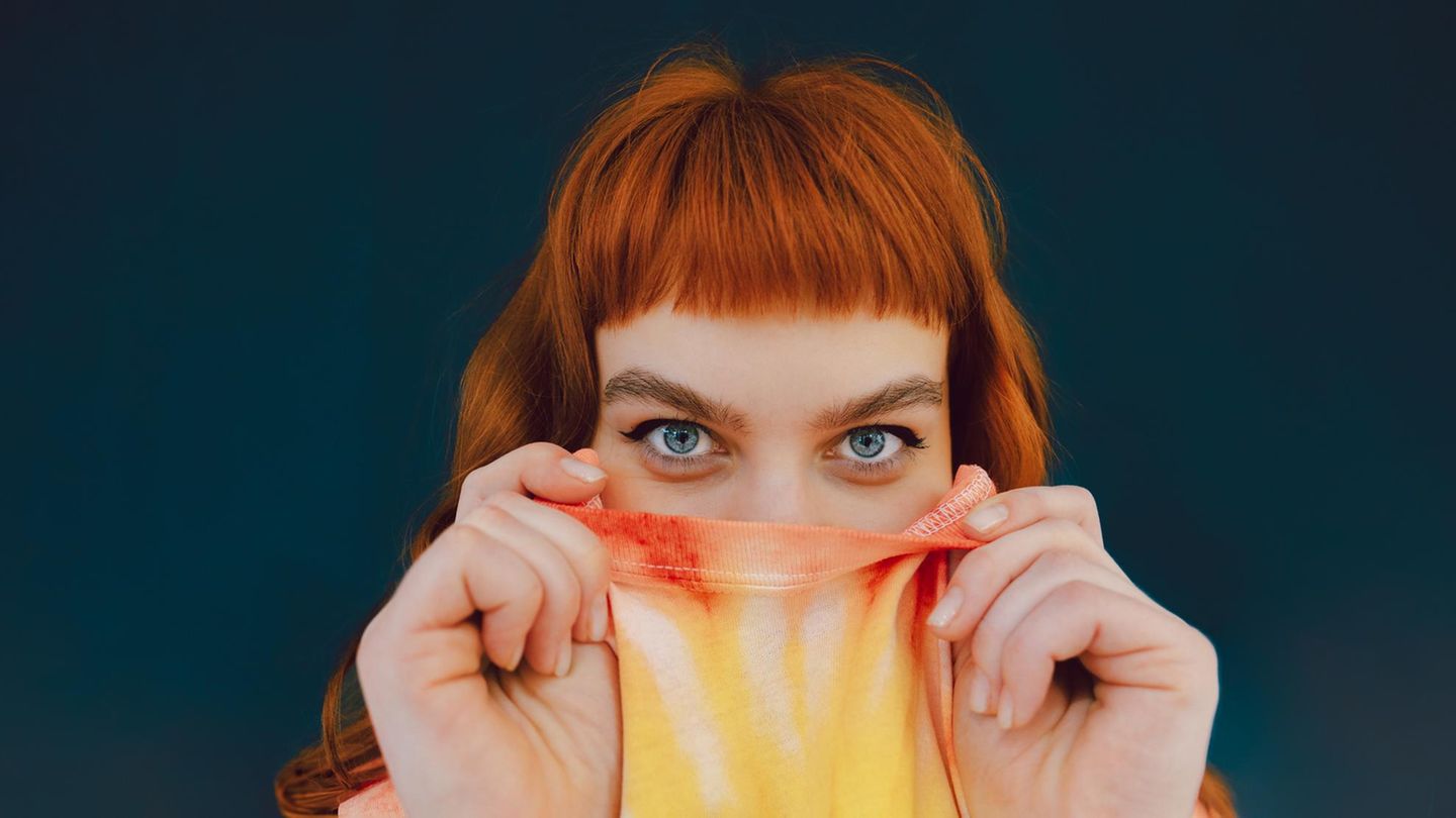 Eine junge Frau versteckt ihre untere Gesichtshälfte in ihrem T-shirt und schaut in die Kamera