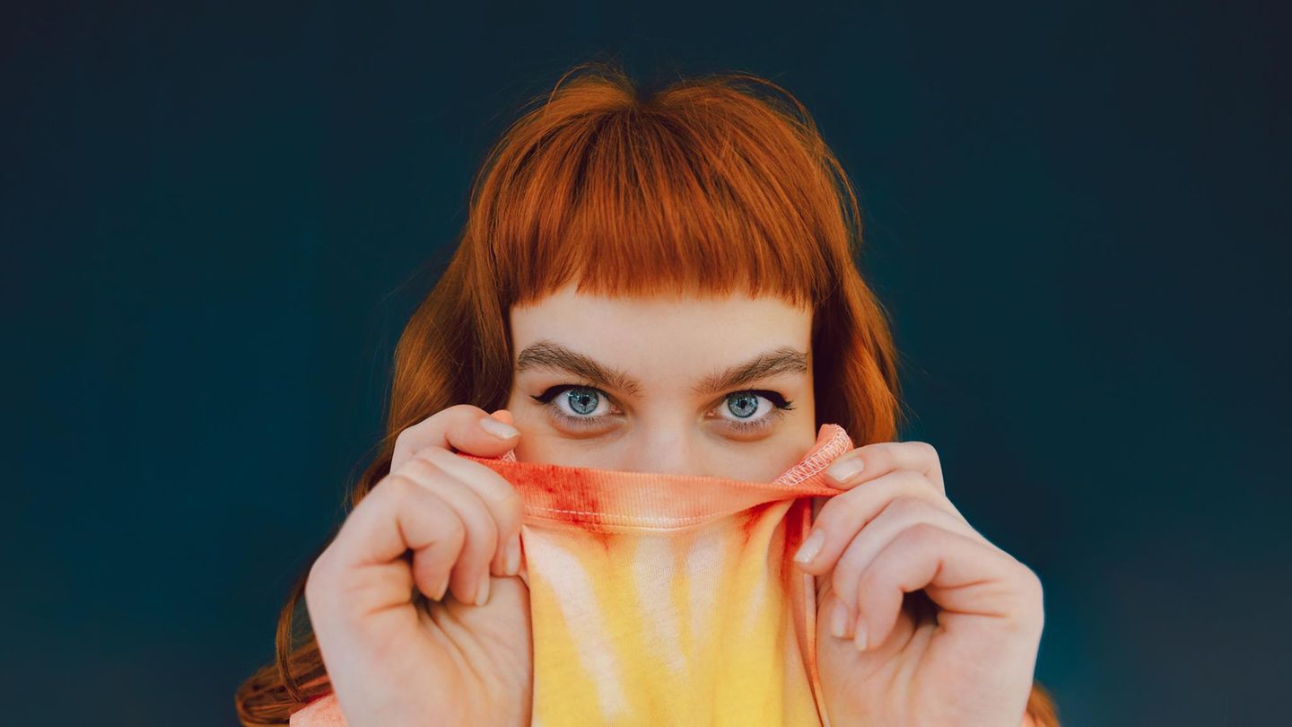 Eine junge Frau versteckt ihre untere Gesichtshälfte in ihrem T-shirt und schaut in die Kamera