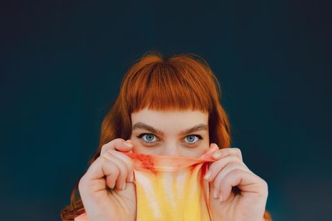 Eine junge Frau versteckt ihre untere Gesichtshälfte in ihrem T-shirt und schaut in die Kamera