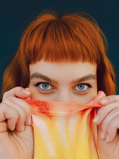 Eine junge Frau versteckt ihre untere Gesichtshälfte in ihrem T-shirt und schaut in die Kamera