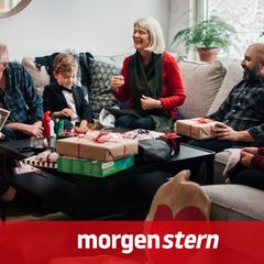 Mehrer Generationen sitzen Weihnachten auf einem Ecksofa, während Kinder Geschenke auspacken