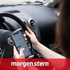 Eine Frau sitzt in einem Auto und tippt mit der linken Hand auf einem Smartphone, während sie mit recht steuert