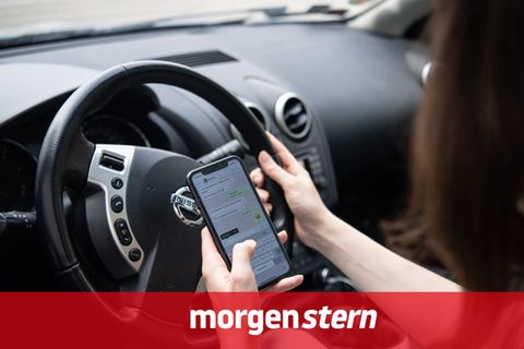 Eine Frau sitzt in einem Auto und tippt mit der linken Hand auf einem Smartphone, während sie mit recht steuert