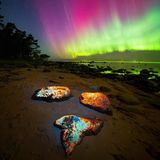 Grün-violettes Polarlicht über einem Strand; darauf leuchtende Steine