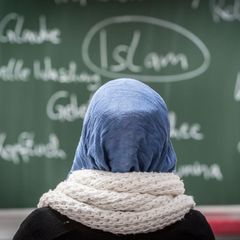 Eine junge Frau mit Kopftuch sitzt in einer Schule vor einer Tafel