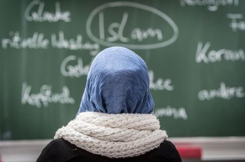 Eine junge Frau mit Kopftuch sitzt in einer Schule vor einer Tafel