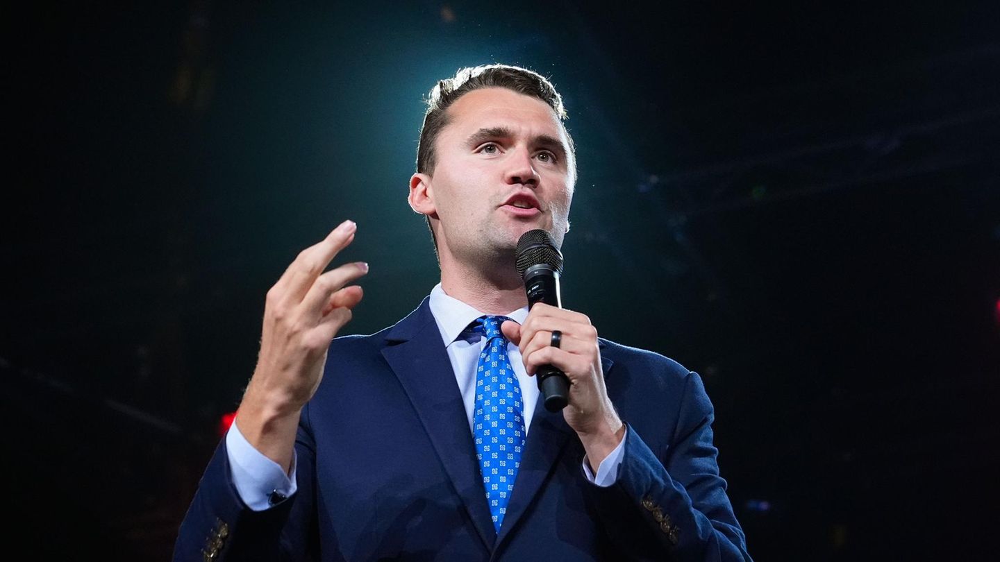 Charlie Kirk steht in Anzug und Krwatte auf einer Bühne und spricht in ein Mikro in seiner linken Hand