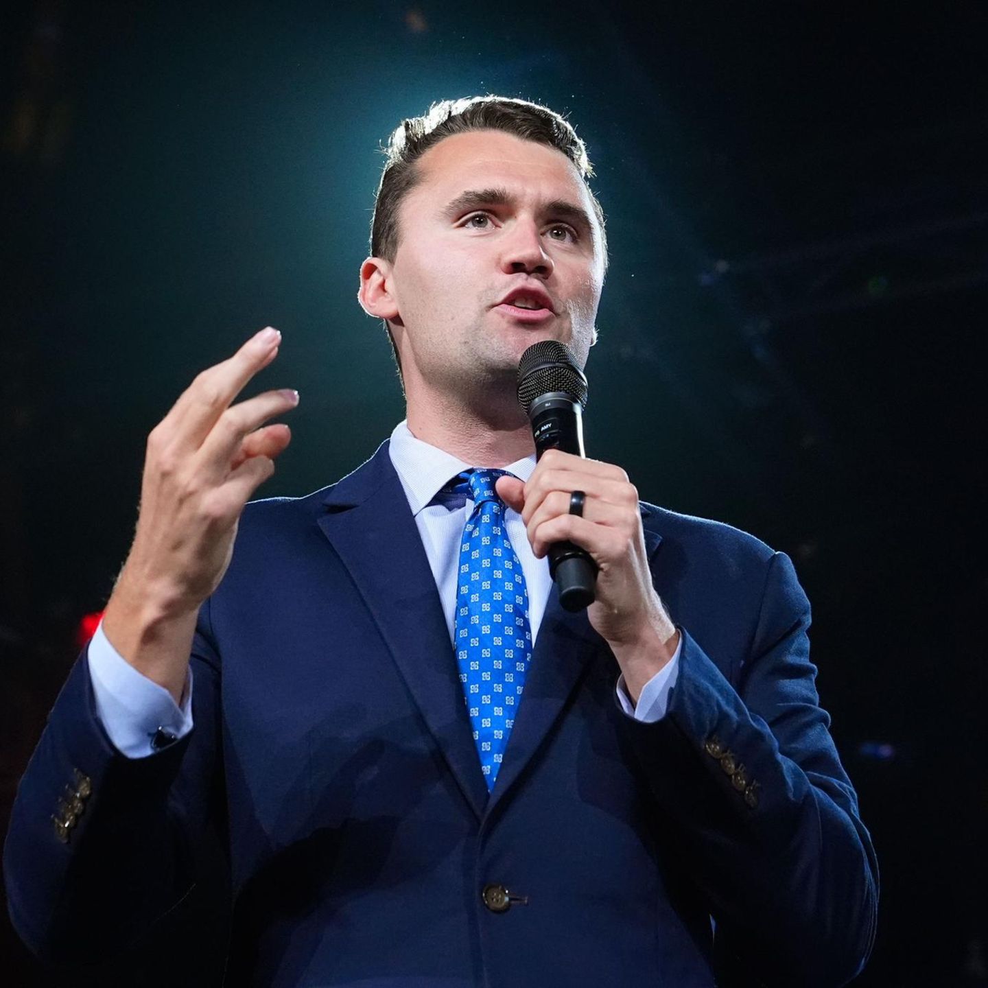 Charlie Kirk steht in Anzug und Krwatte auf einer Bühne und spricht in ein Mikro in seiner linken Hand