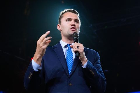 Charlie Kirk steht in Anzug und Krwatte auf einer Bühne und spricht in ein Mikro in seiner linken Hand