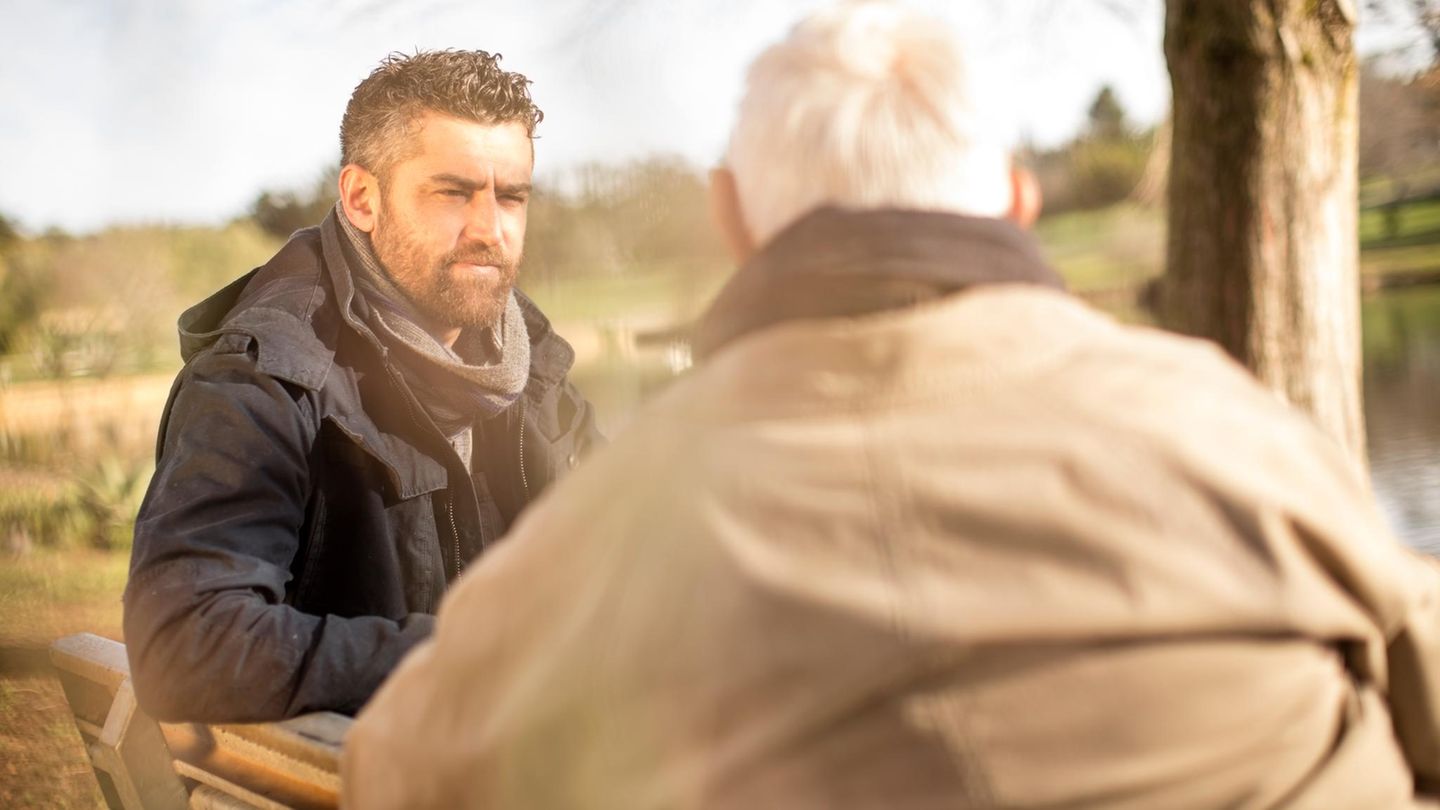 Ein mittelalter Mann mit Bart sitzt mit einem Senioren auf einer Bank im Grünen, die beiden diskutieren – die Rente?