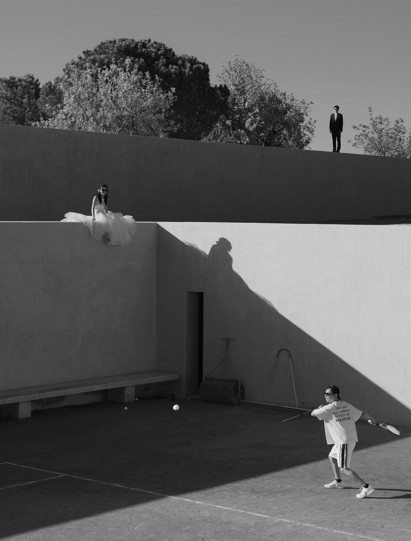Eine Person spielt Tennis, während im Hintergrund zwei Menschen in Hochzeitskleidung stehen und sitzen