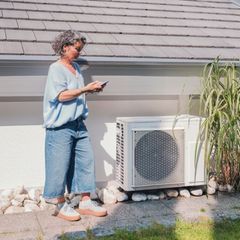 Wärmepumpenstrom-Vergleich: Eine Frau checkt ihr Handy neben ihrer Wärmepumpe